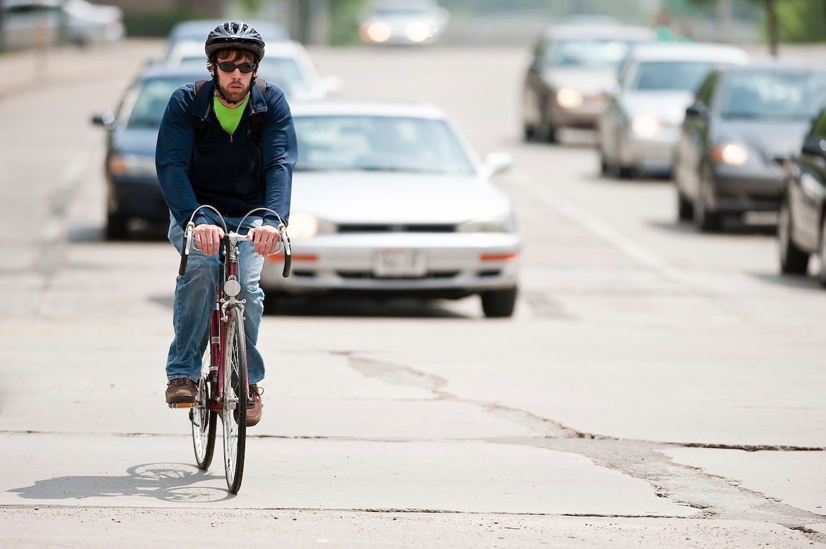 Man with a helmet is riding a bike along a road. Two cars are slightly behind him.
