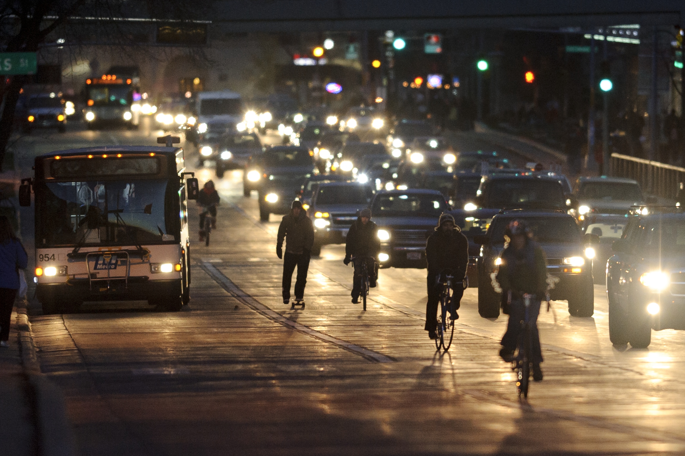 A skateboarder and bicyclists travel in the bike lane along University Avenue at nightfall against a backdrop of vehicle lights and rush hour-traffic.