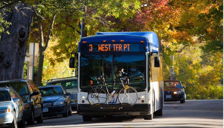 A blue and white Madison Metro Transit bus with a bike on the front rack is driving along a tree and car-lined street.