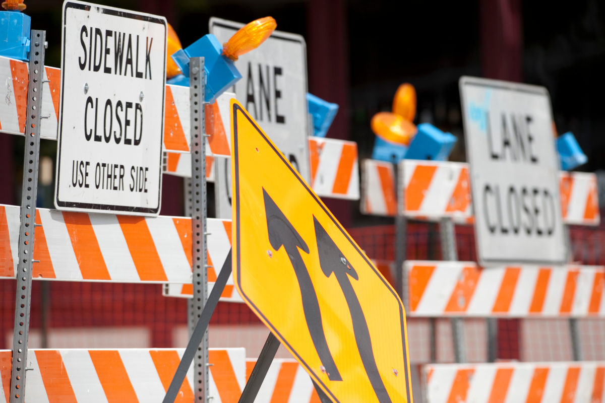 Picture of an area blocked by construction barricades and other traffic management signs. Focus is on two signs noting lane changes, with orange barricades seen in the background with "Sidewalk closed" and "Lane closed" signs.