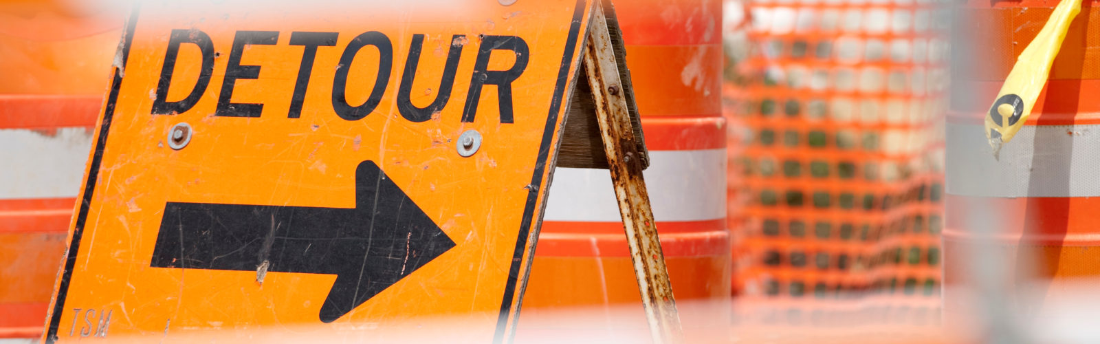 Picture of an area blocked by construction barricades and other traffic management signs. Focus is on a bright orange sign stating "Detour" with an arrow pointing to the right.