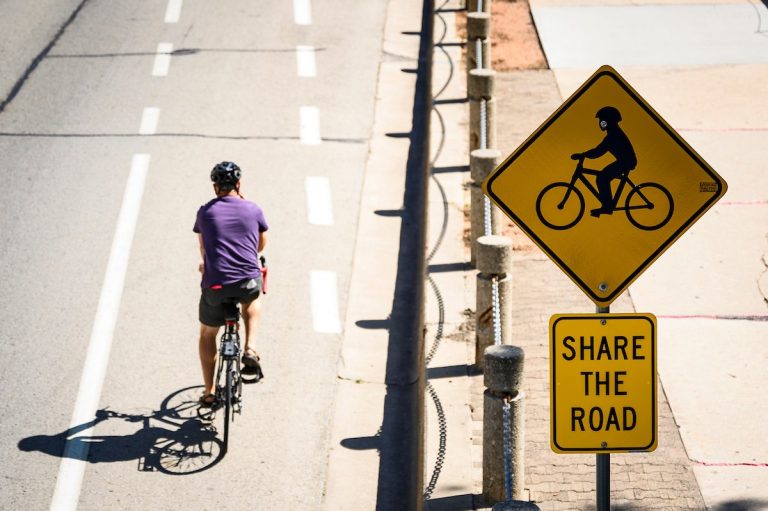 Image of a biker in a purple t-shirt and black shorts wearing a helmet and using a protected bike lane. To the right is a yellow road sign showing a biker with a helmet, another sign below it reading "Share the Road."