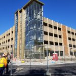 Wide shot of the nearly complete new parking facility. Glass has been installed on the stairwell column. Fencing surround the site and two construction workers in safety gear are seen on the left.