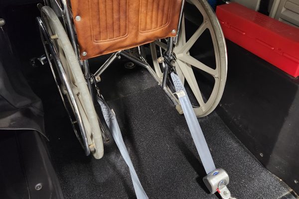 Photo of a wheelchair inside of the rear passenger area of a Riteway accessible shuttle. The photo shows a closeup of the vehicle's rear securements attached to the two rear wheels of the wheelchair. The securements are part of a system which keeps the wheelchair in place while the vehicle is in motion.